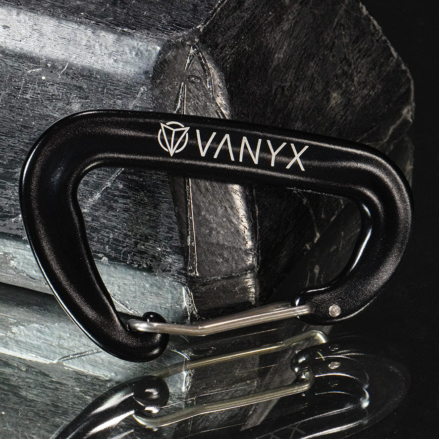 Schwarzer Vanyx Karabiner aus Aluminium mit Logo – liegend auf Stein